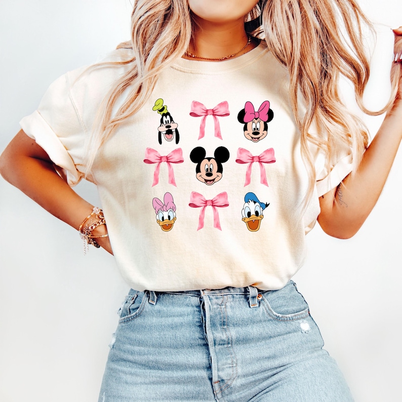 May include: A white t-shirt with a graphic print of various Disney characters, including Mickey Mouse, Minnie Mouse, Donald Duck, and Goofy. Each character is wearing a pink bow.