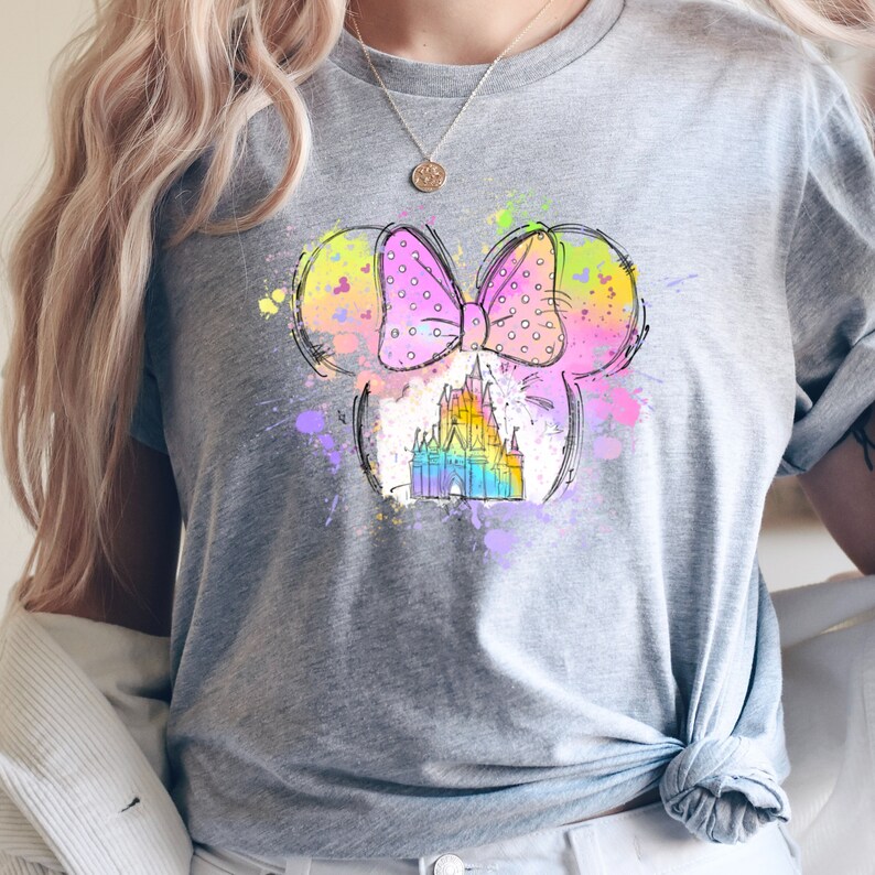 Puede incluir: Una camiseta gris con un gr&aacute;fico colorido de una cabeza de Mickey Mouse con un lazo y un castillo dentro. El gr&aacute;fico tiene un efecto acuarela con colores rosa, azul, amarillo y verde.