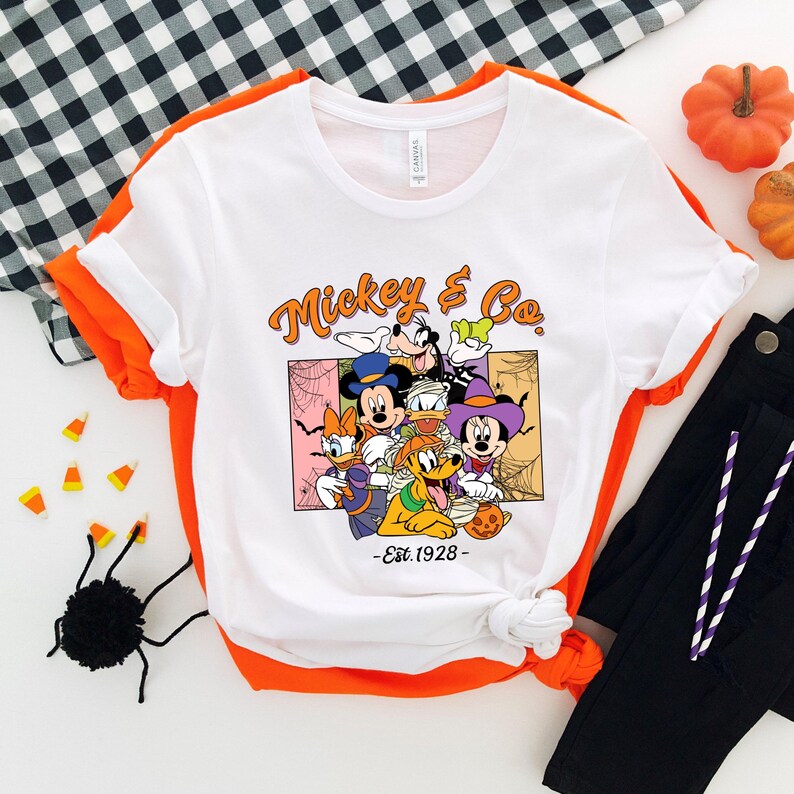 以下が含まれることがあります： ミッキー＆Co.のキャラクターが仮装したハロウィーンをテーマにしたグラフィックがプリントされた白いTシャツ。デザインにはミッキーマウス、ドナルドダック、グーフィー、ミニーマウスが含まれています。「Mickey & Co. - Est. 1928」の文字も含まれています。