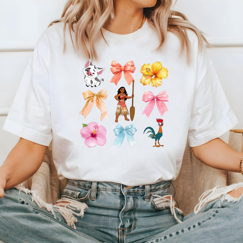 Pu&ograve; includere: Maglietta bianca con una stampa grafica colorata che presenta un maiale dei cartoni animati, una donna con una pagaia, un gallo, fiori e fiocchi.