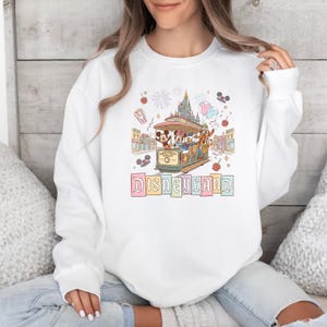 May include: White sweatshirt featuring a colorful Disneyland graphic. The design includes Mickey Mouse, Minnie Mouse, Donald Duck, and Goofy riding a trolley, with a castle and fireworks in the background. The word "Disneyland" is written in pastel colors.