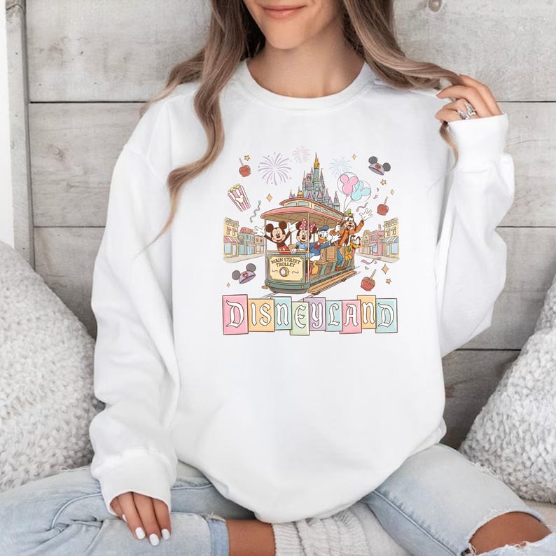 May include: White sweatshirt featuring a colorful Disneyland graphic. The design includes Mickey Mouse, Minnie Mouse, Donald Duck, and Goofy riding a trolley, with a castle and fireworks in the background. The word "Disneyland" is written in pastel colors.