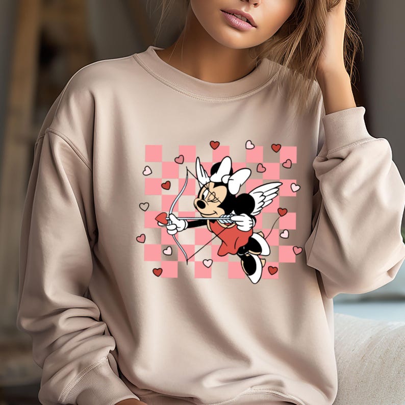 Pode incluir: Uma camisola bege com um padr&atilde;o xadrez rosa e branco que apresenta uma imagem de desenho animado de Minnie Mouse vestida como Cupido com um arco e flecha. O design inclui cora&ccedil;&otilde;es cor-de-rosa.