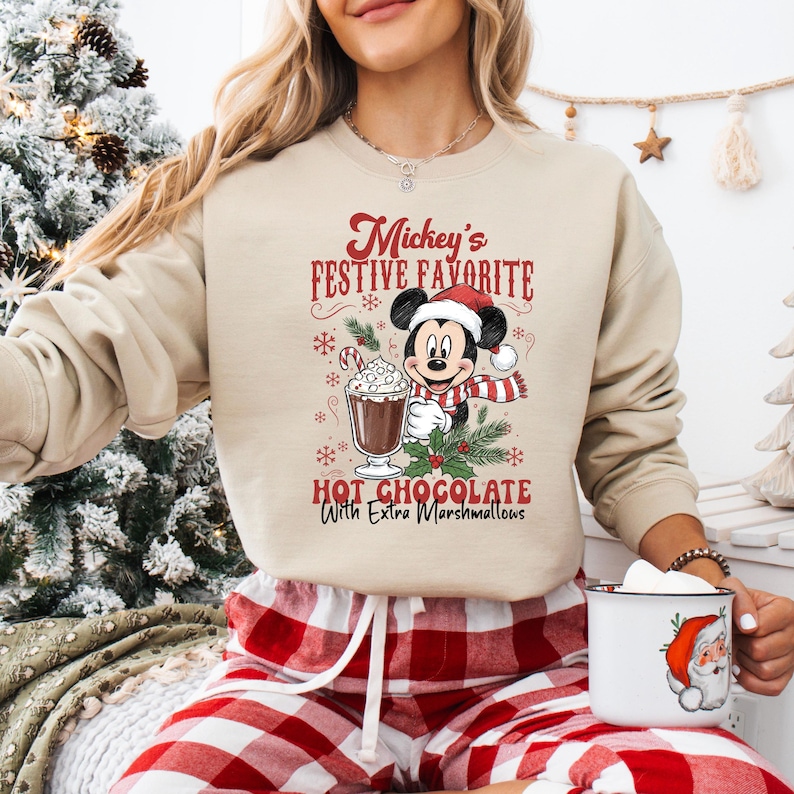 May include: Beige sweatshirt with a graphic of Mickey Mouse in a Santa hat and scarf, holding holly. Text reads "Mickey's Festive Favorite Hot Chocolate With Extra Marshmallows". The person is wearing red and white plaid pajama pants.