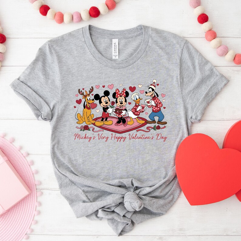 May include: A light gray t-shirt featuring a Valentine's Day graphic with Disney characters. Mickey, Minnie, Donald, Goofy, and Pluto are depicted with hearts and roses. The text "Mickey's Very Happy Valentine's Day" is printed below the characters.