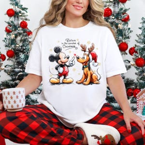 May include: White t-shirt featuring Mickey Mouse and Pluto in a Christmas design. Mickey Mouse is wearing red shorts and Pluto has reindeer antlers and a Santa hat. The text "Believe in the wonder of Christmas" is above the characters.