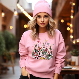 May include: Pink sweatshirt with a Christmas-themed design featuring Mickey Mouse, Minnie Mouse, Donald Duck, and Goofy standing in front of Christmas trees with balloons.