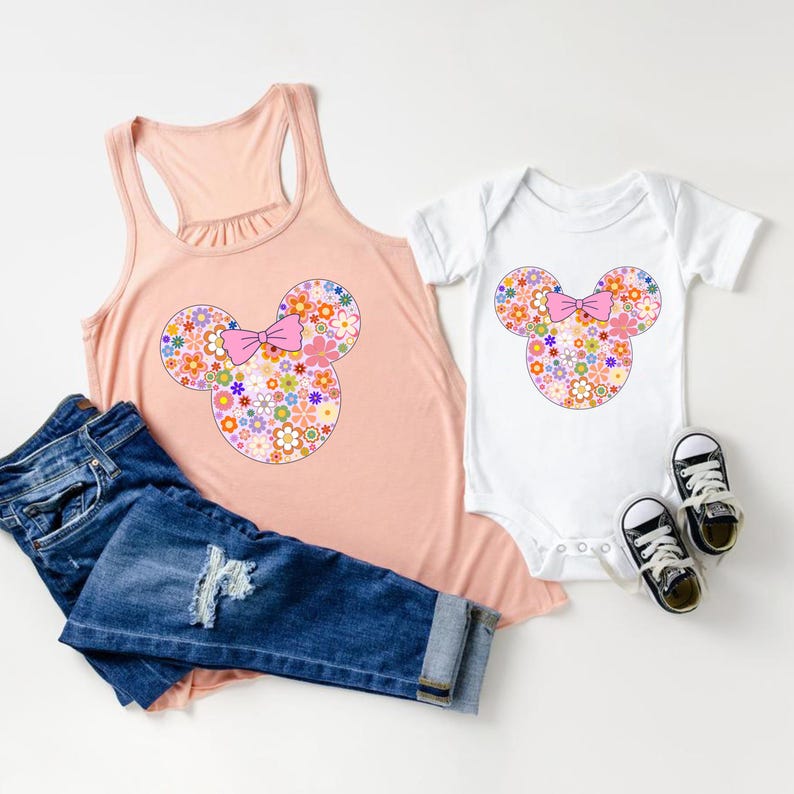 May include: A peach-colored tank top and a white baby onesie, both featuring a floral Mickey Mouse design with a pink bow. Dark blue denim jeans and black sneakers complete the ensemble.