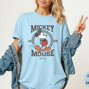 May include: Light blue t-shirt featuring a distressed graphic of Mickey Mouse. The design includes the text "Mickey Mouse" and the years "1928". The shirt is a Comfort Colors Chambray.