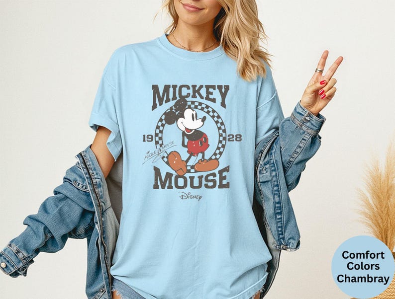 May include: Light blue t-shirt featuring a distressed graphic of Mickey Mouse. The design includes the text "Mickey Mouse" and the years "1928". The shirt is a Comfort Colors Chambray.