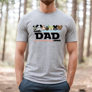 May include: Gray t-shirt with a graphic featuring cartoon character heads and the word "DAD" in black. A red lightsaber is below the text. The shirt has short sleeves and a crew neck.