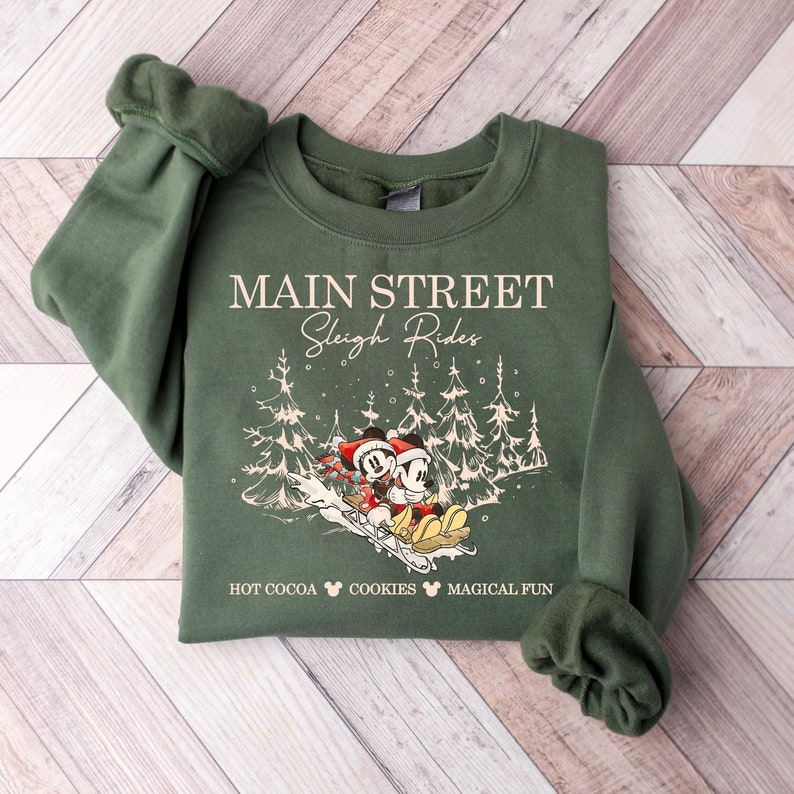 May include: Green sweatshirt with the text "Main Street Sleigh Rides" and a graphic of Mickey and Minnie Mouse sledding. The sweatshirt also has the words "Hot Cocoa", "Cookies", and "Magical Fun".