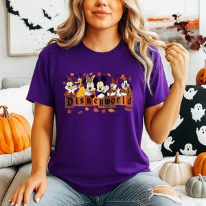 May include: A purple t-shirt featuring Disney characters, including Mickey and Minnie Mouse, Donald and Daisy Duck, and Goofy. The word "Disneyland" is printed in brown letters. Autumn leaves and pumpkins are in the background.