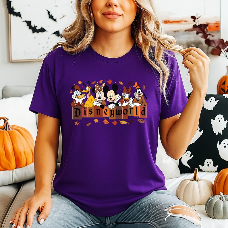 May include: A purple t-shirt featuring Disney characters, including Mickey and Minnie Mouse, Donald and Daisy Duck, and Goofy. The word "Disneyland" is printed in brown letters. Autumn leaves and pumpkins are in the background.