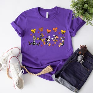 May include: A purple t-shirt with a Halloween design featuring cartoon characters holding Mickey Mouse-shaped balloons. The shirt is styled with denim shorts and white trainers. A tassel and sunglasses are also visible.