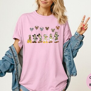 K&ouml;nnte beinhalten: Hellrosa T-Shirt mit einer Cartoon-Illustration von Disney-Figuren, die gemusterte Ballons halten. Die Figuren sind Pluto, Mickey, Minnie, Daisy und Donald. Das Shirt hat auch die Worte "Comfort Colors Blossom" in einem rosa Kreis.