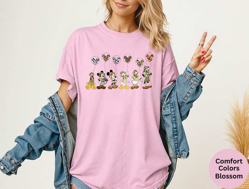 K&ouml;nnte beinhalten: Hellrosa T-Shirt mit einer Cartoon-Illustration von Disney-Figuren, die gemusterte Ballons halten. Die Figuren sind Pluto, Mickey, Minnie, Daisy und Donald. Das Shirt hat auch die Worte "Comfort Colors Blossom" in einem rosa Kreis.
