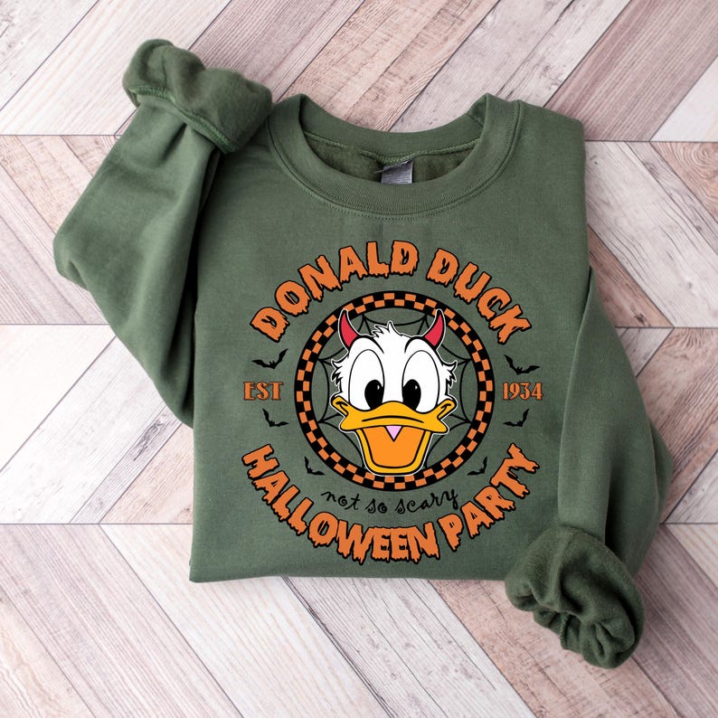 May include: Green sweatshirt featuring a circular graphic of Donald Duck with devil horns. The graphic reads "Donald Duck Est 1934 Halloween Party not so scary".