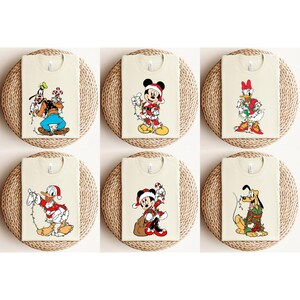 May include: Six cream-colored t-shirts featuring Disney characters in Christmas attire. Goofy, Mickey Mouse, Donald Duck, Minnie Mouse, and Pluto are shown with festive lights and Santa hats. Each shirt is displayed on a woven circular mat.