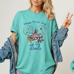 May include: Seafoam green t-shirt with a graphic featuring Disney characters celebrating the 4th of July. The design includes fireworks, a castle, and the text "Happy 4th of July est. 1776 David".