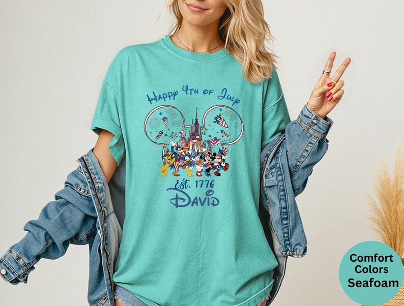 May include: Seafoam green t-shirt with a graphic featuring Disney characters celebrating the 4th of July. The design includes fireworks, a castle, and the text "Happy 4th of July est. 1776 David".