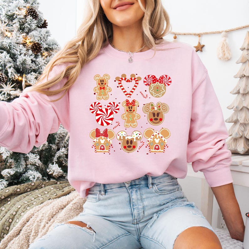 May include: Pink Christmas sweater with a gingerbread character design featuring Mickey Mouse, Minnie Mouse, and other characters. The characters are decorated with red and white candy canes, sprinkles, and icing.