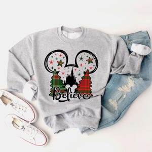 Puede incluir: Sudadera gris con un dise&ntilde;o de Mickey Mouse en cuadros rojos y verdes con un castillo y estrellas. El texto "Believe" est&aacute; impreso debajo del dise&ntilde;o.