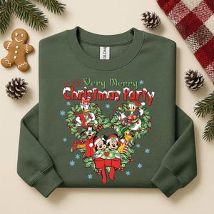 Op de afbeelding: Groene sweatshirt met een kerstontwerp met Disney-personages. De afbeelding bevat Mickey en Minnie Mouse, Donald en Daisy Duck en Goofy, met de tekst "Very Merry Christmas Party."