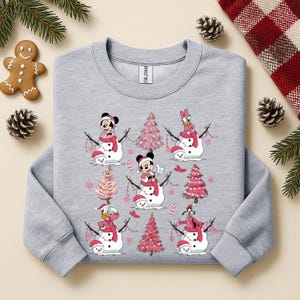 May include: A light grey sweatshirt with a festive design featuring cartoon snowmen and pink Christmas trees. The snowmen have character faces and wear red hats and scarves. A gingerbread man and pinecones are in the background.