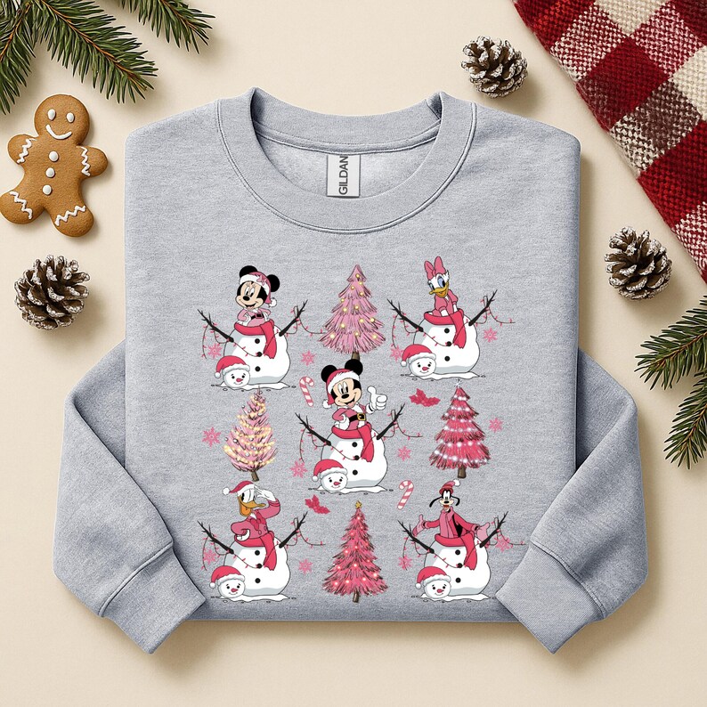 May include: A light grey sweatshirt with a festive design featuring cartoon snowmen and pink Christmas trees. The snowmen have character faces and wear red hats and scarves. A gingerbread man and pinecones are in the background.