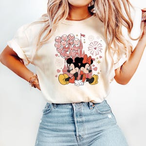 May include: A white t-shirt with a pink and red graphic of Mickey Mouse and Minnie Mouse in front of a pink castle with pink balloons and fireworks. The text "Love" is on one of the balloons.