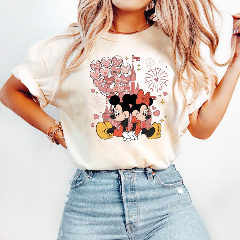 May include: A white t-shirt with a pink and red graphic of Mickey Mouse and Minnie Mouse in front of a pink castle with pink balloons and fireworks. The text "Love" is on one of the balloons.