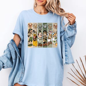 May include: Light blue t-shirt with a graphic print of Mickey Mouse, Minnie Mouse, Donald Duck, Goofy, and Pluto dressed in safari outfits. The characters are holding balloons with animal print designs.