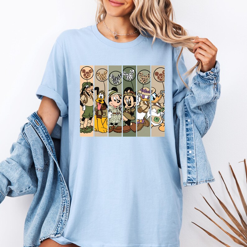 May include: Light blue t-shirt with a graphic print of Mickey Mouse, Minnie Mouse, Donald Duck, Goofy, and Pluto dressed in safari outfits. The characters are holding balloons with animal print designs.