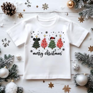 May include: White t-shirt featuring four Christmas trees with mouse ears, decorated with stars and snowflakes. The text "merry christmas" is written in a script font below the trees. Festive holiday apparel.