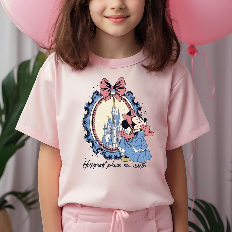 May include: Pink short-sleeved t-shirt with a graphic of Minnie Mouse in a blue dress standing in front of Cinderella's Castle. The graphic is framed with a blue and gold ornate frame with a pink bow on top. The text "Happiest place on earth" is printed below the graphic.