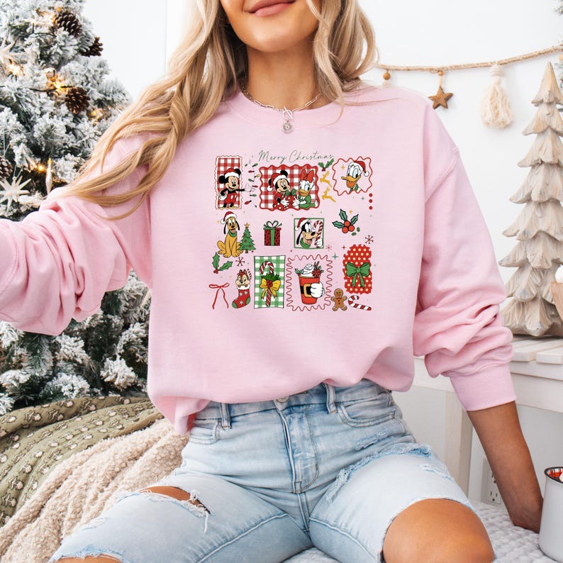 May include: A light pink sweatshirt featuring a festive Christmas design with cartoon characters and the words "Merry Christmas." The design includes Mickey Mouse, Minnie Mouse, Donald Duck, and other holiday elements.