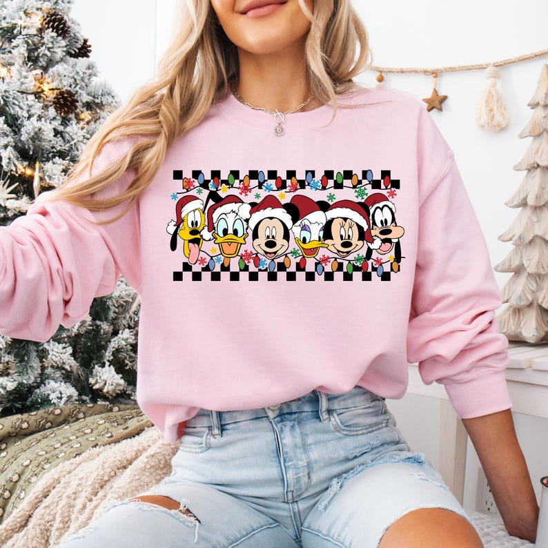 May include: Pink Christmas sweatshirt with a checkered pattern and a row of Disney characters wearing Santa hats. The characters include Mickey Mouse, Minnie Mouse, Donald Duck, Goofy, and Pluto.