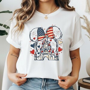 May include: White t-shirt featuring a patriotic design. The graphic includes a castle, Mickey Mouse ears with an American flag pattern, hearts, and fireworks. The design is in red, white, blue, and yellow.