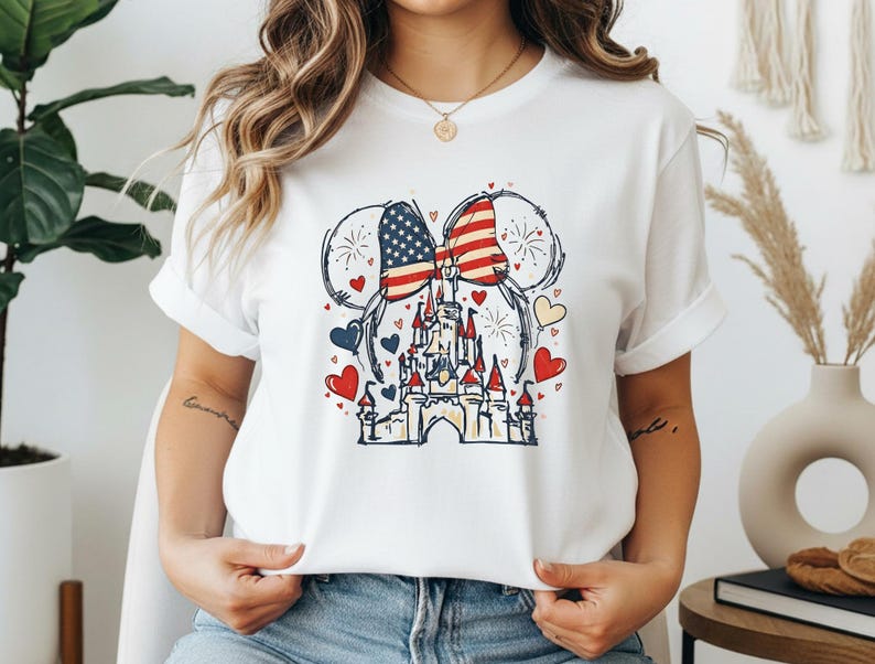 May include: White t-shirt featuring a patriotic design. The graphic includes a castle, Mickey Mouse ears with an American flag pattern, hearts, and fireworks. The design is in red, white, blue, and yellow.