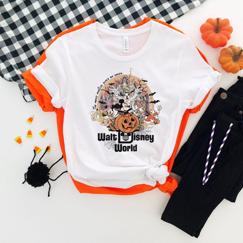 Puede incluir: Camiseta blanca con un dise&ntilde;o gr&aacute;fico de Halloween con Mickey Mouse, una calabaza tallada y un castillo. El texto "Walt Disney World" est&aacute; debajo. La camiseta se combina con pantalones negros y detalles en naranja.