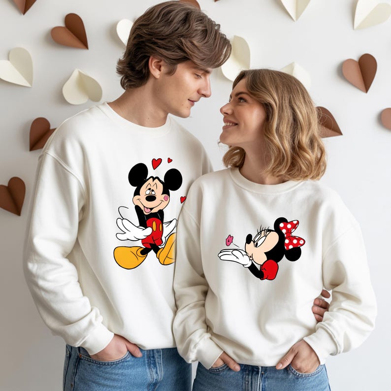 May include: Matching cream-colored sweatshirts featuring Mickey and Minnie Mouse designs. The Mickey Mouse sweatshirt has a graphic of Mickey with hearts, while the Minnie Mouse sweatshirt shows Minnie blowing a kiss. Both sweatshirts are worn by a couple.