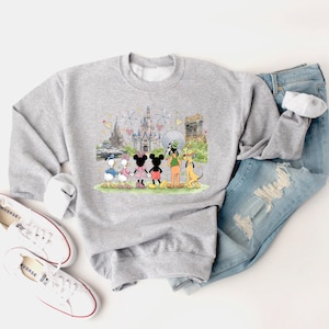May include: A light gray sweatshirt featuring a colorful graphic of Disney characters, including Mickey and Minnie Mouse, Donald and Daisy Duck, Goofy, and Pluto, in front of various park landmarks. The sweatshirt is paired with blue jeans and white sneakers.