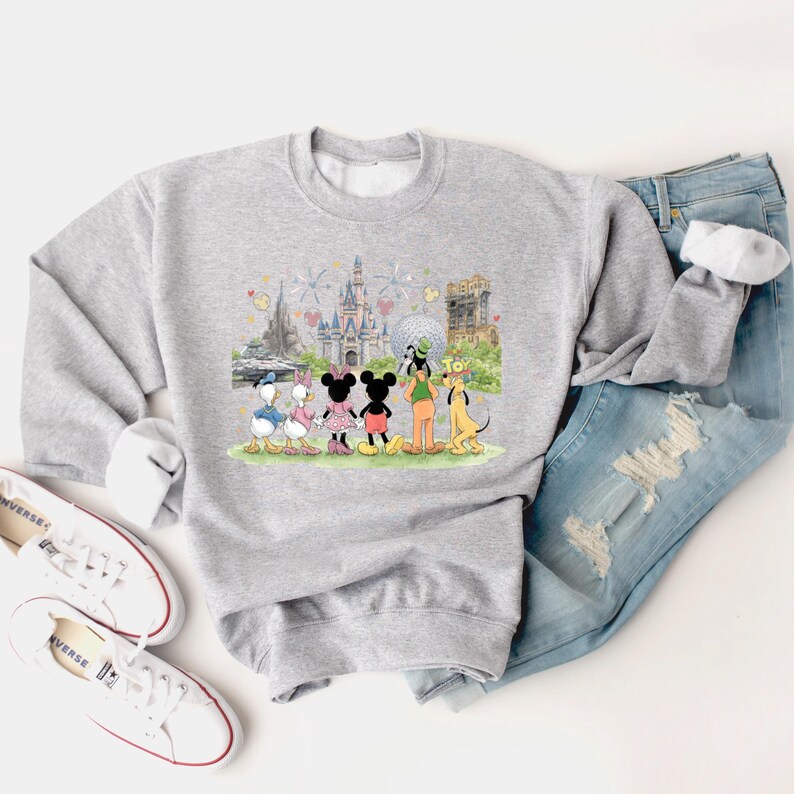 May include: A light gray sweatshirt featuring a colorful graphic of Disney characters, including Mickey and Minnie Mouse, Donald and Daisy Duck, Goofy, and Pluto, in front of various park landmarks. The sweatshirt is paired with blue jeans and white sneakers.
