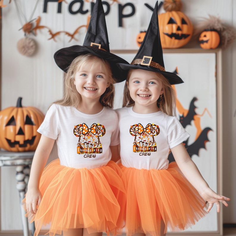 Puede incluir: Dos ni&ntilde;as peque&ntilde;as con camisetas blancas con un dise&ntilde;o de Acci&oacute;n de Gracias, tut&uacute;s naranjas y sombreros de bruja negros. Las camisetas tienen el texto "Thanksgiving Crew". Las calabazas y las decoraciones de Halloween est&aacute;n en el fondo.