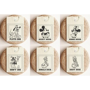 May include: Six cream-colored t-shirts featuring black line drawings of Disney characters: Pluto Dog, Mickey Mouse, Minnie Mouse, Goofy Dog, Donald Duck, and Daisy Duck. Each design is framed with a black border and character name.