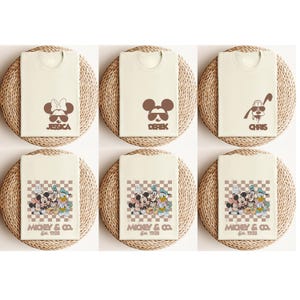 May include: Cream-colored t-shirts with brown cartoon character designs. Some shirts feature character faces with names like "Jessica", "Derek", and "Chris". Other shirts display a group of characters with the text "Mickey & Co. Est. 1928".
