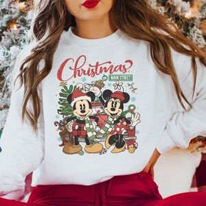 Op de afbeelding: Witte trui met een kerstontwerp met Mickey en Minnie Mouse voor een rode auto. De tekst op de trui luidt "Christmas on Main Street".