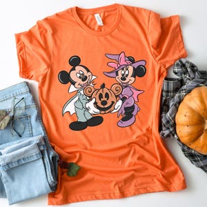 May include: Orange t-shirt with a graphic of Mickey Mouse and Minnie Mouse dressed in Halloween costumes. Mickey is a vampire and Minnie is a witch. They are holding a jack-o'-lantern.