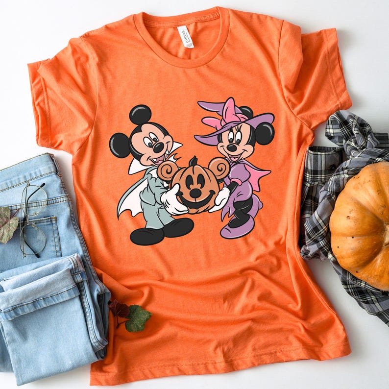 May include: Orange t-shirt with a graphic of Mickey Mouse and Minnie Mouse dressed in Halloween costumes. Mickey is a vampire and Minnie is a witch. They are holding a jack-o'-lantern.